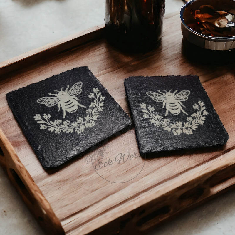 Two black slate coasters with a floral design engraved on them, placed on a wooden tray.