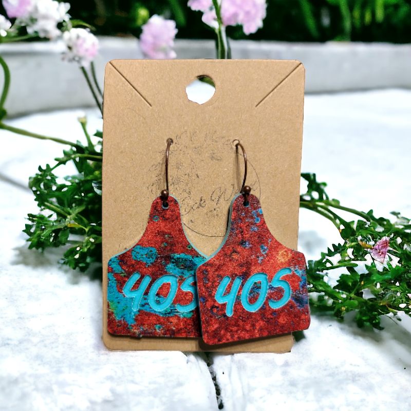 A pair of custom copper and teal earrings with '405' engraved, displayed on a cardboard holder with a floral background. 