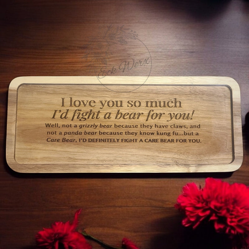 A wooden trinket tray with the phrase 'I love you so much I'd fight a bear for you' laser-engraved on it, displayed on a wooden surface with red flowers around it.
