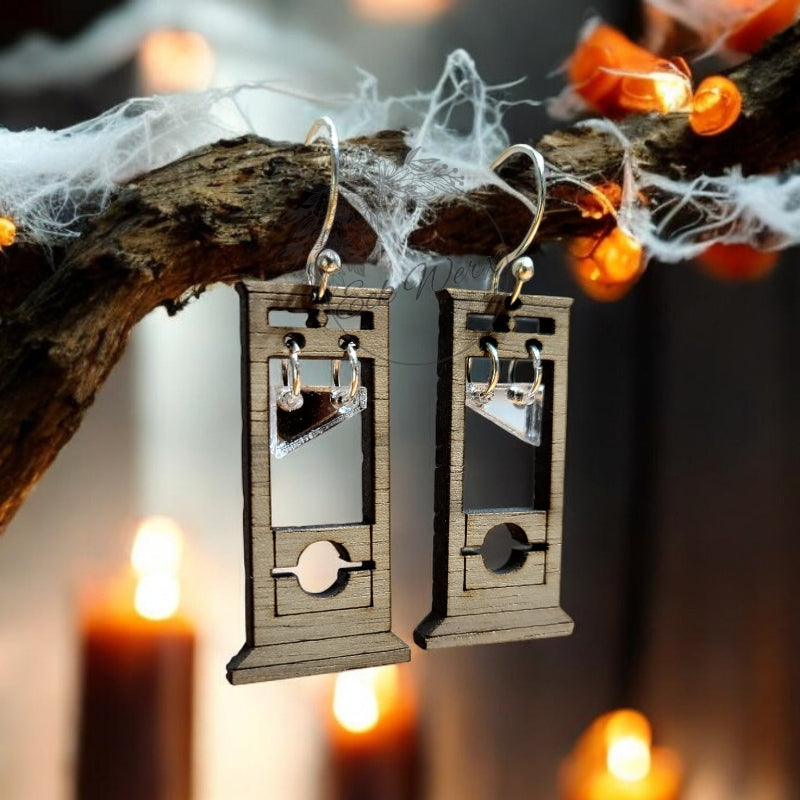 Wooden guillotine earrings hanging on a branch with spider webs and blurred candles in the background. EckWerx LLC