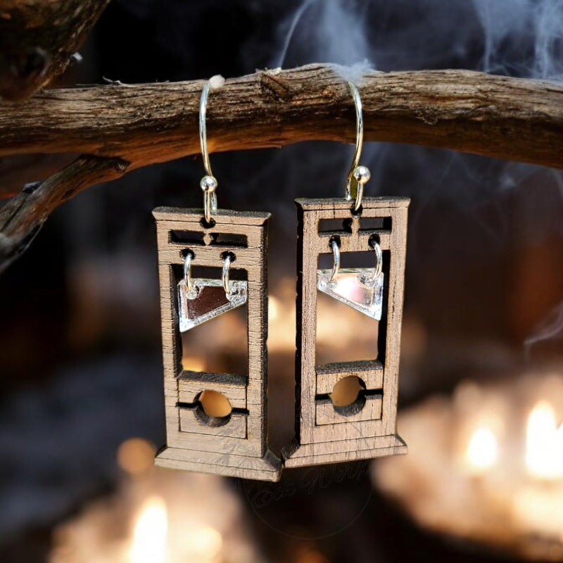A pair of handcrafted wooden guillotine earrings displayed on a branch with a background of lit candles and a spooky Halloween setting.