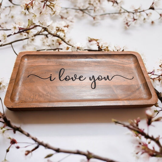 A wooden tray with the engraved text 'I love you' in a flowing script, displayed on a table with floral backdrop.